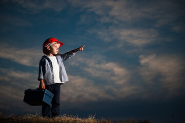 job child labor concept of one little worker kid in orange red helmet pointing at copy space on dark blue cloudy sky background