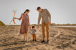 © Lena May - Happy family walking on sandy beach of river. Father, mother holding baby son on hands and playing with kite.