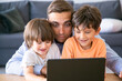 © Mangostar - Middle-aged dad hugging sons and watching movie on laptop. Happy little boys sitting at table with father, looking at screen and smiling. Fatherhood, childhood and digital technology concept