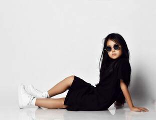 Cool asian kid girl in stylish black dress and round sunglasses sitting on floor side to camera with one her knee up