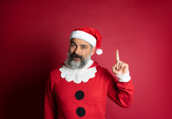 Bearded man wearing christmas hat pointing finger up looking at camera on red background