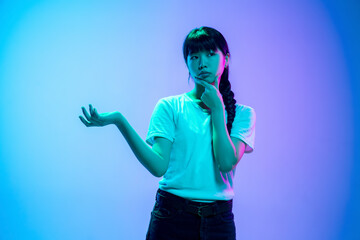 Wall Mural - Thoughtful, choosing. Young asian woman's portrait on gradient blue-purple studio background in neon light. Concept of youth, human emotions, facial expression, sales, ad. Beautiful brunette model.