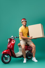 cheerful delivery man in helmet holding big carton box near scooter on blue