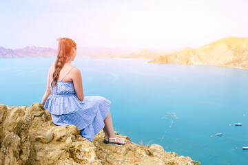 Girl in a blue dress sits on the mountain and admires the seascape. The traveler looks at the mountains and the sea. Hiking, adventure and active tourism