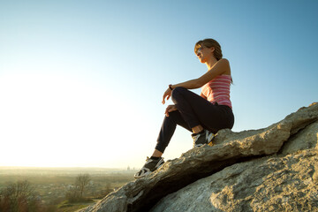Wall Mural - Woman hiker sitting on a steep big rock enjoying warm summer day. Young female climber resting during sports activity in nature. Active recreation in nature concept.