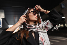 Elegant Young Pretty Beautiful Woman In Stylish Dark Sunglasses In A Black Coat Puts A Luxurious Silk Scarf On Head Outdoors. Beautiful Girl Fashion Model Relaxes On The Road In The City. Sexy Lady.