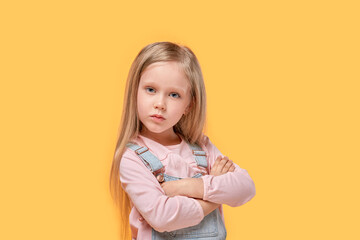 Blonde baby girl in casual clothes looks at the camera. Yellow isolated background.