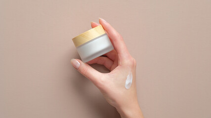 female hand holding jar of cosmetic cream on pastel beige background. woman applying moisturizer cre