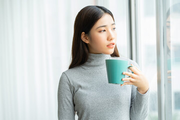 Wall Mural - Asian woman drinking coffee by the window during break
