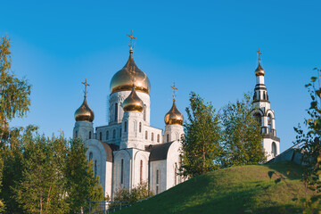 Orthodox Church made of white stone with Golden domes among green trees against a blue sky. The topic is faith, religion