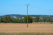 A Wooden Electric Pole Standing In The Middle Of A Plowed Field, In The Background Hills With Trees.