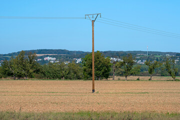 A wooden electric pole standing in the middle of a plowed field, in the background hills with trees.