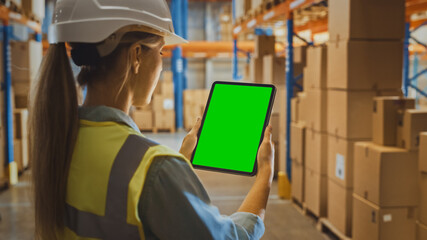 Wall Mural - Professional Female Worker Wearing Hard Hat Uses Digital Tablet Computer with Green Chroma Key Screen in Portrait Mode in the Retail Warehouse full of Shelves with Goods. Logistics and Distribution
