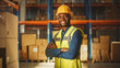 © Gorodenkoff - Handsome and Happy Professional Worker Wearing Safety Vest and Hard Hat Smiling with Crossed Arms on Camera. In the Background Big Warehouse with Shelves full of Delivery Goods. Medium Portrait