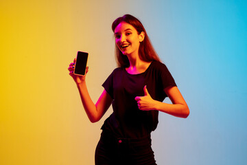 Wall Mural - Showing blank phone screen. Young caucasian woman's portrait on gradient blue-yellow studio background in neon light. Concept of youth, human emotions, facial expression, sales, ad. Beautiful model.