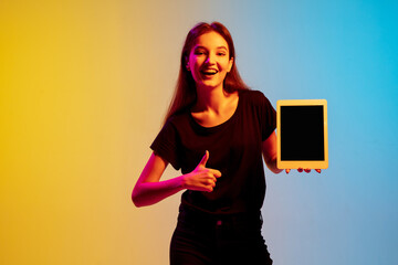 Wall Mural - Showing blank tablet screen. Young caucasian woman's portrait on gradient blue-yellow studio background in neon light. Concept of youth, human emotions, facial expression, sales, ad. Beautiful model.