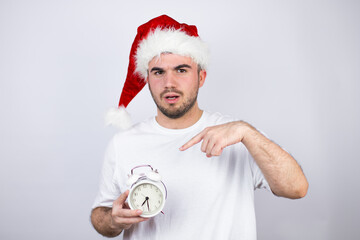 Young handsome man wearing a Santa hat over white background pointing, scared and holding a clock