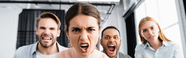 Angry businesswoman looking at camera near multiethnic colleagues on blurred background in office, banner