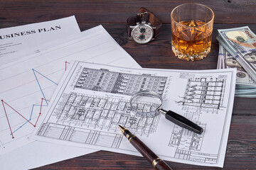 Business papers with building layout and pen. Glass magnifier with money, clock and glass of whiskey.