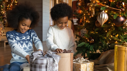 Wall Mural - Close up smiling little African American siblings, cute girl and boy wearing sweaters unwrapping Christmas gifts, sitting on floor near decorated festive tree at home, celebrating New Year