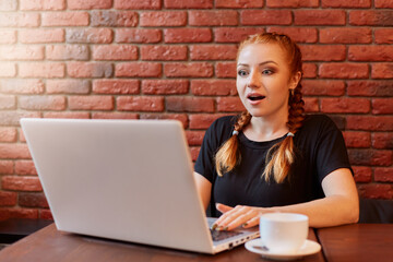 Surprised astonished young woman with red hair and two pigtails, sitting with open mouth, being in amazement, shocked by unexpected news, poses isolated on brick wall, drinks coffee while working.