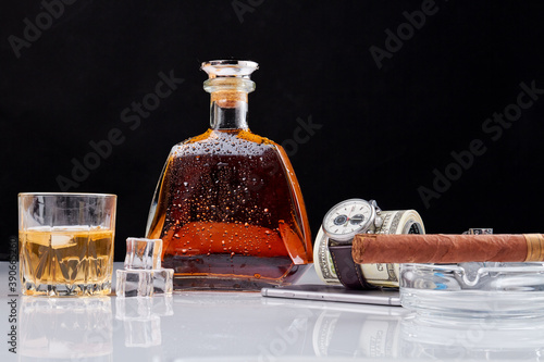 Composition of attributes of success. Bottle of brandy with class cup and cigar. Isolated on black background.