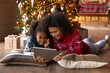 Smiling African American woman with daughter using tablet, having fun with device, lying on pillows on warm floor near Christmas tree at home, happy family shopping online, choosing gifts