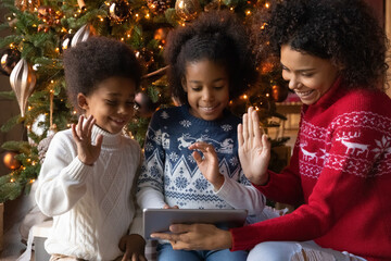 Close up happy African American family making video call to relatives on Christmas, sitting near festive tree at home, smiling mother with two kids using tablet, chatting online, waving hands