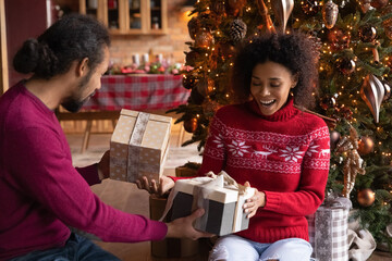 Overjoyed young African American man and woman giving Christmas gift boxes, presents, sitting on floor near decorated festive tree, family celebrating New Year at home, enjoying winter holidays