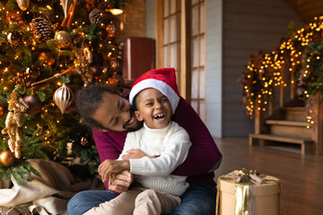 Wall Mural - Overjoyed African American man hugging adorable little son wearing festive red cap, family celebrating Christmas, opening gifts, sitting on warm floor at home near tree, enjoying winter holidays