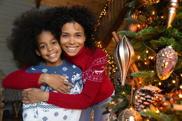 Wall Mural - Head shot portrait smiling young African American mother and adorable little daughter wearing sweaters standing near decorated pine tree at home, celebrating Christmas, hugging and looking at camera