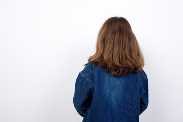 The back view of a Little caucasian girl with beautiful blue eyes wearing denim jacket standing over isolated white background. Studio Shoot.