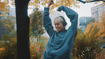 Young blond short haired woman with hoodie listening to the music on heaset and dancing in the city park in autumn. High quality photo