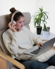 Poster - Vertical shot of a young European woman using a laptop with headset sitting on the chair at home