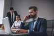 © zinkevych - Man with smartphone at laptop, colleagues nearby