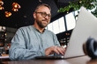 © dusanpetkovic1 - Middle aged bearded smiling freelancer with eyeglasses sitting in a restaurant and using laptop to type report. Middle aged persons can be excellent freelancers.