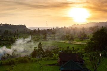 Wall Mural - Scenic sunset over ricefields meadow smoke and rural village in the background at Bali, Indonesia