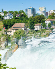 Wall Mural - Panorama des cascades du Rhin ou Rheinfall en Suisse par une journée ensoleillée avec un ciel bleu avec les rochers et un village et les embruns du fleuve.