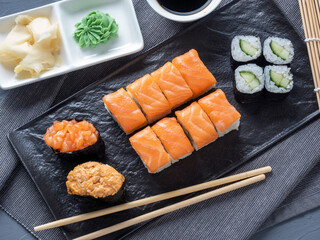 a variety of rolls and sushi gunkan nested on a black plate. next to it are bamboo wasabi sticks and sauce. Top view, flat lay. traditional Japanese cuisine