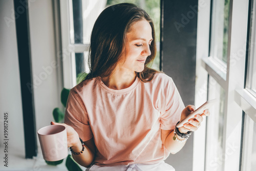 Fotomural Attractive young woman in pink t-shirt with cup of coffee surfs internet with mo