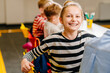 © Iryna - Smiling girl sitting at desk in class room and looking at camera. Portrait of young black schoolgirl studying with classmates in background. Happy smiling pupil writing on notebook.