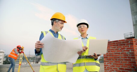 Wall Mural - Caucasian male and female builders, architectors with draft plan of building and tablet device talking on constructing site. Man and woman constructors working together at construction. Disscussion.