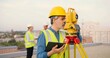 Caucasian man topographer in casque measuring angle with total station on building roof. Male builder constructor doing topographic measures and using tablet device. Geodesic concept. Device computer.
