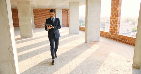 Wall Mural - Caucasian male real-estate agent in glasses, suit and tie walking in building on construction phase, using tablet device and tapping or scrolling. Handsome businessman investor choosing mortgage.