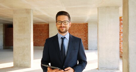 Wall Mural - Portrait of Caucasian handsome young man in glasses, suit and tie standing at construction site indoor. Male real-estate agent with tablet device in hands. Zooming in. Dolly shot. Businessman.