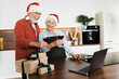 © Kateryna - Celebrating Christmas online. Happy elderly man and woman in Christmas hats stand in the kitchen with wine and gifts and meet the new year by video link with family