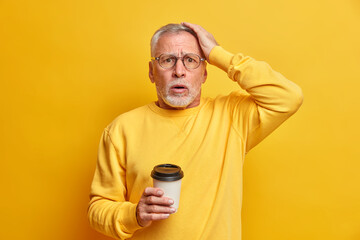 Concerned embarrassed bearded aged man keeps hand on head and stares stupefied at camera drinks takeaway coffee dressed in casual jumper isolated over yellow background cannot believe in failure