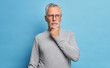 © wayhome.studio  - Old age and thoughts concept. Serious grey haired man holds chin and concentrated with pensive expression somewhere dressed in casual turtleneck isolated over blue background thinks over problems