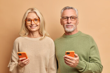 Wall Mural - Horizontal shot of elderly couple use modern technologies hold smart phones reads text messages connected to wireless internet wear casual jumpers isolated over brown background. Retired pensioners