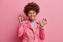 Photo Of Funny Dark Skinned Young Woman Makes Cat Claws Clenches Teeth Makes Roar And Looks Playfully At Camera Gestures Actively Dressed In Fashionable Jacket Isolated Over Pink Background.